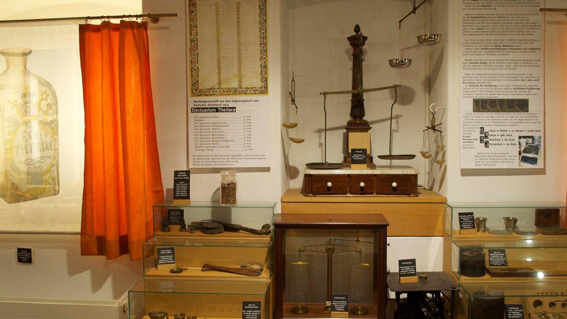 Ein Ausstellungsraum mit historischen Objekten wie Waagen und Messgeräten. Im Hintergrund sind informative Texte und ein roter Vorhang zu sehen. | © Theriak Museum