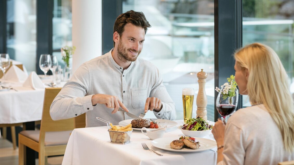 Ein elegantes Restaurant mit einem Mann und einer Frau, die am Tisch sitzen. Der Mann schneidet ein Steak, während die Frau ein Glas Wein hält. | © Eisenberger