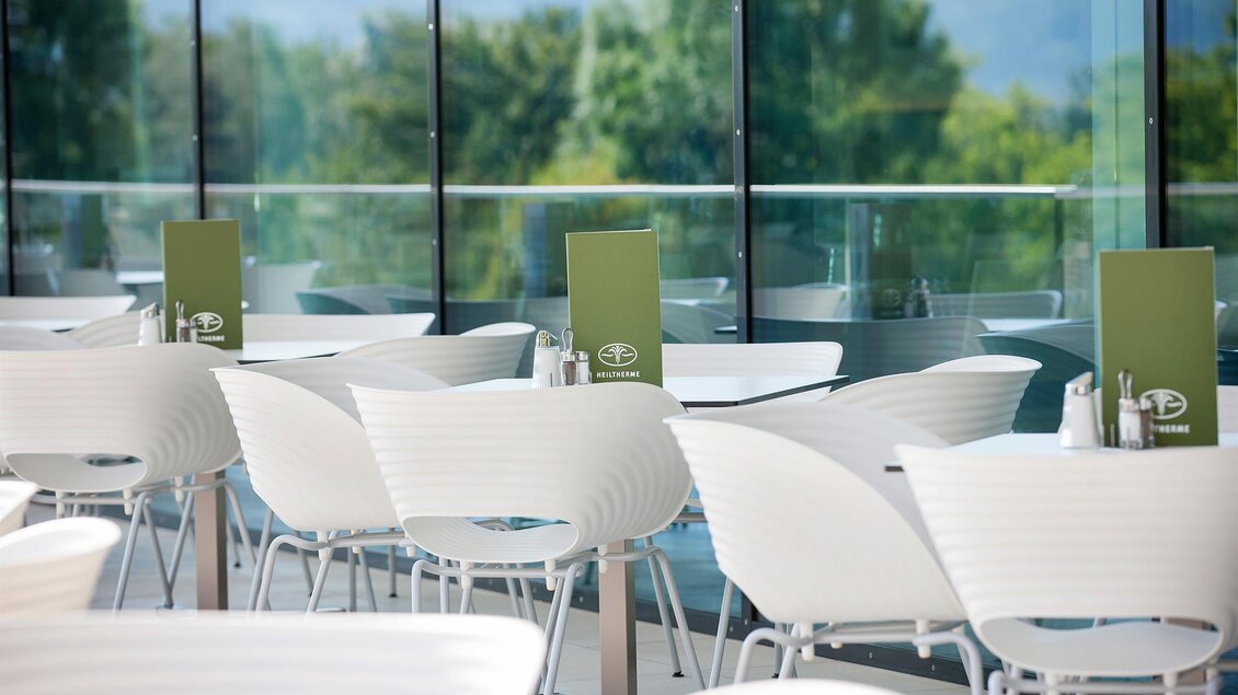 Ein modernes Restaurant mit weißen Stühlen und Tischen. Durch große Fenster ist die grüne Landschaft im Hintergrund sichtbar. | © Eisenberger