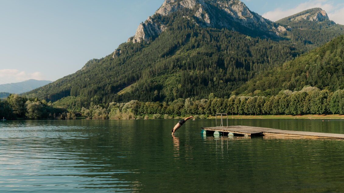 Ein ruhiger See umgeben von grünen Bergen. Ein Holzsteg führt ins Wasser. | © (c) Region Graz - studio draussen