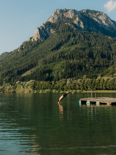 A quiet lake surrounded by green mountains. A wooden jetty leads into the water. | © (c) Region Graz - studio draussen