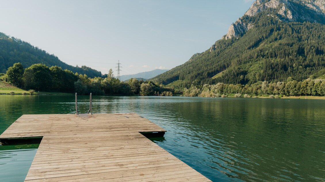 Ein Holzsteg führt zu einem ruhigen See, umgeben von grünen Bergen. Der Himmel ist klar und die Landschaft ist friedlich. | © (c) Region Graz - studio draussen