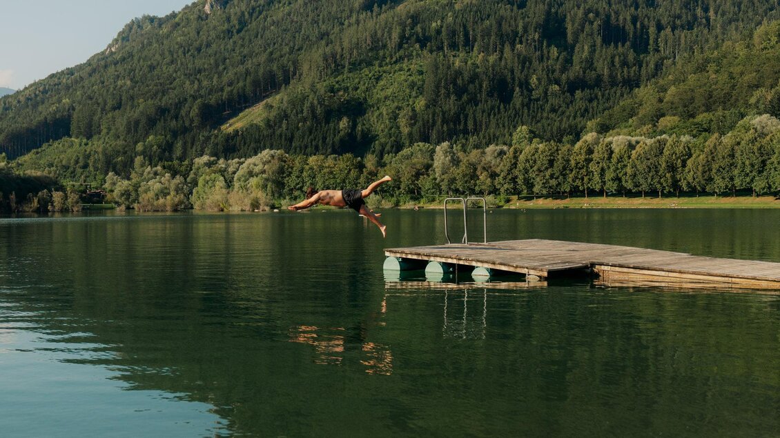 Ein malerischer See mit einem Holzsteg und einer Berglandschaft im Hintergrund. Die ruhige Wasseroberfläche spiegelt die Natur wider. | © (c) Region Graz - studio draussen
