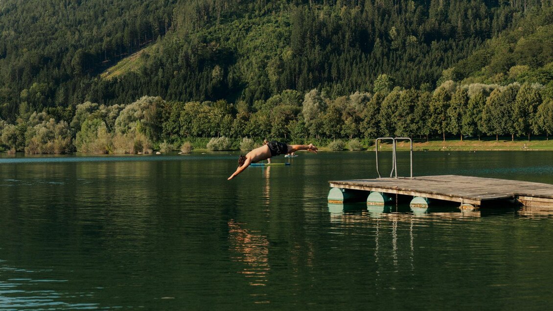 Ein klarer See mit einem Holzsteg und einem Sportler, der ins Wasser springt. Im Hintergrund sind grüne Hügel und Berge zu sehen. | © (c) Region Graz - studio draussen