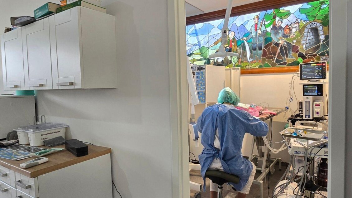 Ein medizinisches Behandlungszimmer mit einem Mitarbeiter in Schutzkleidung. Im Hintergrund ist ein buntes Fenster mit einer ländlichen Szene zu sehen. | © Stephanie Bor