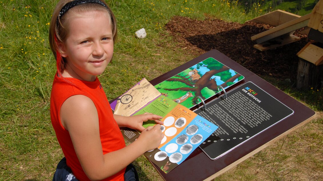 Ein Mädchen sitzt an einem Tisch und hält ein Heft mit verschiedenen Bildern. Es scheint, als würde sie etwas über die Natur lernen. | © Erwin Petz