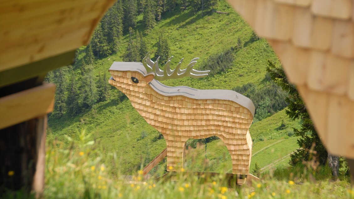 Eine große, hölzerne Statue eines Hirsches steht auf einer grünen Wiese. Im Hintergrund sind weitere Bäume und Berglandschaften sichtbar. | © Erwin Petz