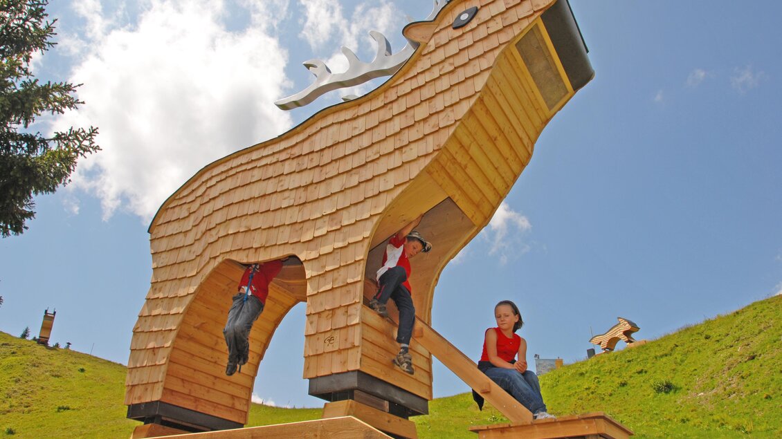 Eine große Holzkonstruktion in Form eines Tieres, die auf einer grünen Wiese steht. Kinder spielen und klettern an der Skulptur. | © Erwin Petz