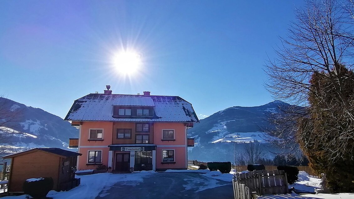 Ein schön gelegenes Haus im Schnee, umgeben von Bergen. Die Sonne scheint strahlend am blauen Himmel. | © Tieschnhof