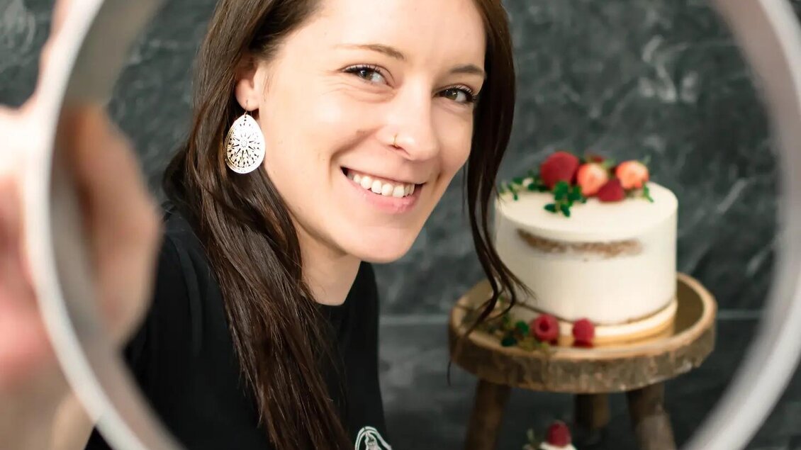 Eine lächelnde Frau hält einen runden Rahmen vor sich. Im Hintergrund steht eine Torte mit Früchten auf einem Tisch. | © Martina Pöltl / Nilepixx by Nicky Hoferfotografie