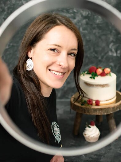 Eine lächelnde Frau hält einen runden Rahmen vor sich. Im Hintergrund steht eine Torte mit Früchten auf einem Tisch. | © Martina Pöltl / Nilepixx by Nicky Hoferfotografie