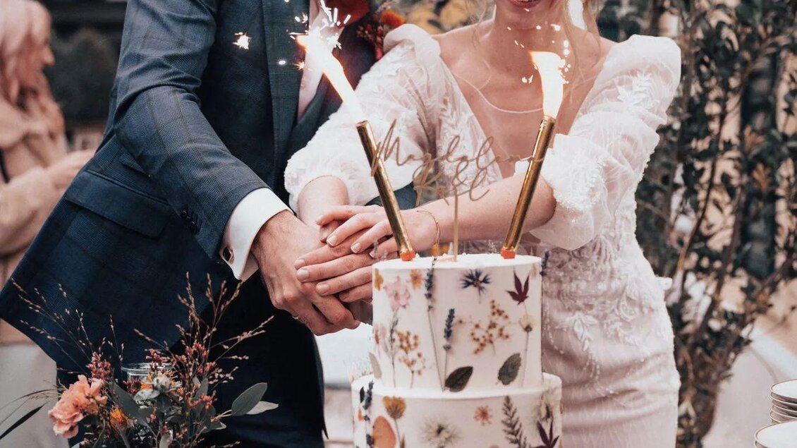 Ein Brautpaar schneidet gemeinsam eine wunderschöne Hochzeitstorte mit Blumendesign. Im Hintergrund sind festlich dekorierte Tables und Gäste zu sehen. | © Martina Pöltl / Nilepixx by Nicky Hoferfotografie