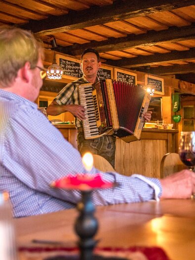A man plays the accordion in a cozy tavern. In the foreground, guests are seated at a table with drinks. | © Tourismusverband Murau