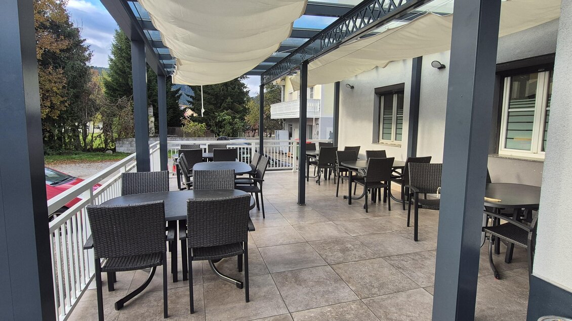 Eine ruhige Terrasse mit Tischen und Stühlen unter einem Sonnenschutz. Im Hintergrund sind Bäume und ein Gebäude sichtbar. | © TV Hochsteiermark