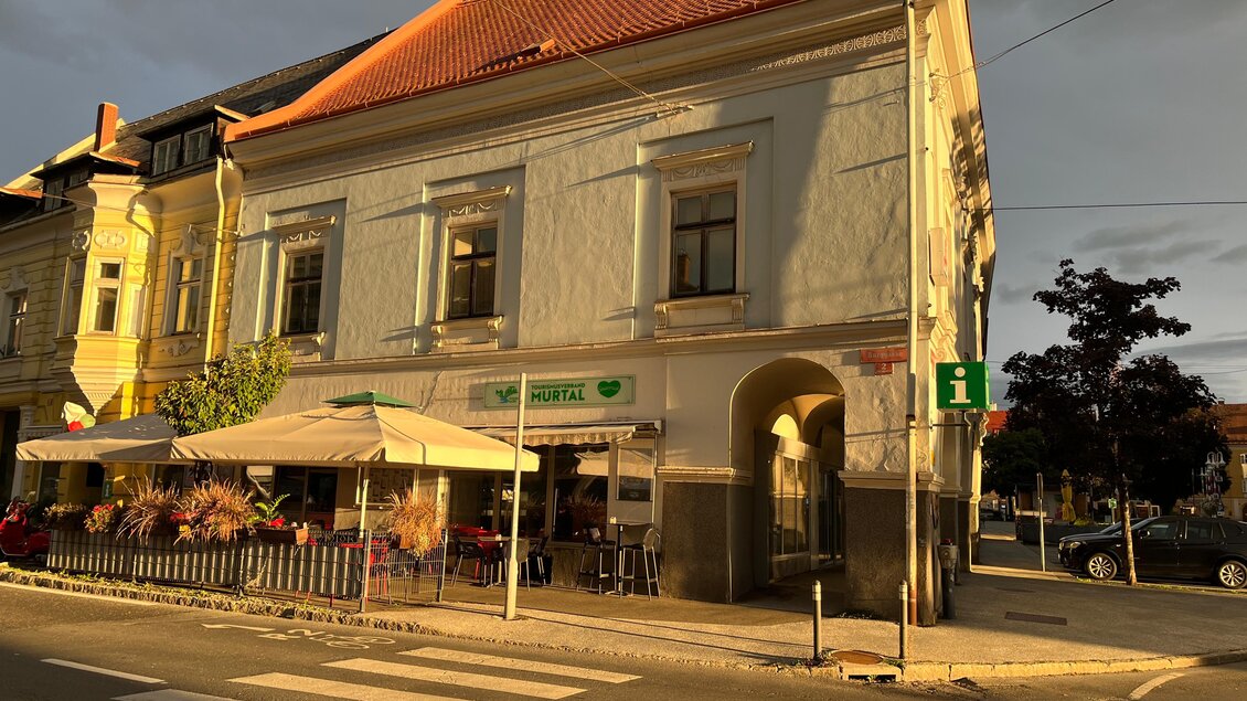Ein historisches Gebäude mit einer terrassenartigen Café-Außenansicht. Die Sonne beleuchtet die Fassade, während die Straßenlaterne und die Umgebung ruhige Stimmung erzeugen. | © Erlebnisregion Murtal