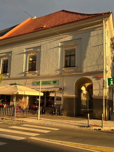 Ein historisches Gebäude mit einer terrassenartigen Café-Außenansicht. Die Sonne beleuchtet die Fassade, während die Straßenlaterne und die Umgebung ruhige Stimmung erzeugen. | © Erlebnisregion Murtal