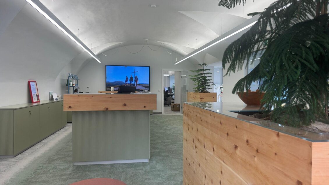 Ein modernes Büro mit Holz- und Pflanzenelementen. Eine große Bildschirmanzeige ist sichtbar und das Design ist hell und einladend. | © Selina Senf