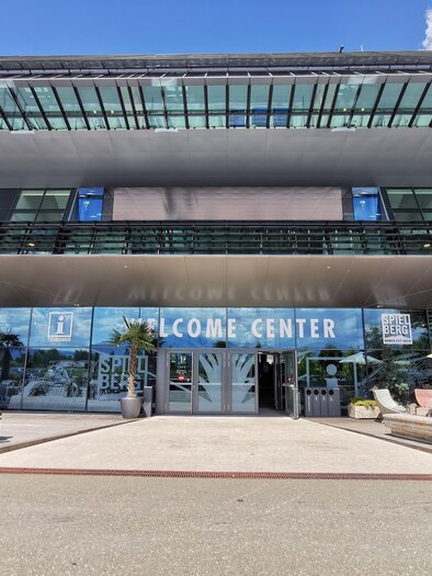 A modern building with the inscription "WELCOME CENTER". In front of the entrance are plants and a table with a sunshade. | © Patrizia Schreibmayr