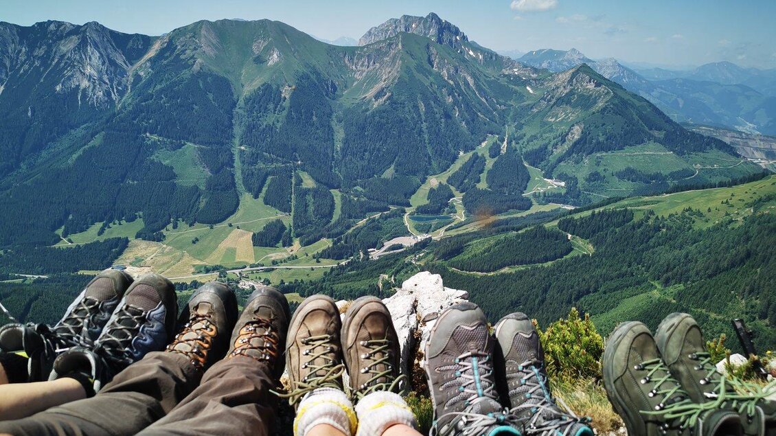 Eine Gruppe von Menschen sitzt auf einem Berggipfel und zeigt ihre Wanderschuhe. Im Hintergrund erstreckt sich eine beeindruckende Berglandschaft mit grünen Wiesen und blauen Himmel. | © TV ERZBERG LEOBEN