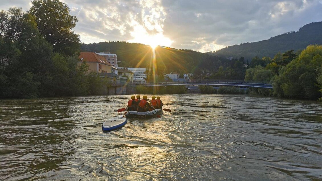 Eine Gruppe von Menschen paddelt in einem Boot auf einem Fluss. Die Sonne geht hinter den Bergen unter und wirft ein warmes Licht auf die Szene. | © LE Bootshaus