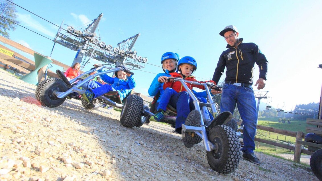 Zwei Kinder sitzen in bunten Go-Karts auf einem Schotterweg. Im Hintergrund sind Seilbahnen und ein blauer Himmel zu sehen. | © Der Wilde Berg