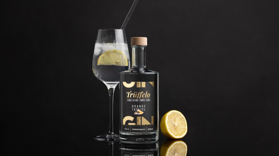 Ein stilvoller Gin mit einer Flasche und einem Glas, gefüllt mit Eis und einer Zitronenscheibe. Der Hintergrund ist schwarz, was die Eleganz der Präsentation unterstreicht. | © True-fellow Handel GmbH