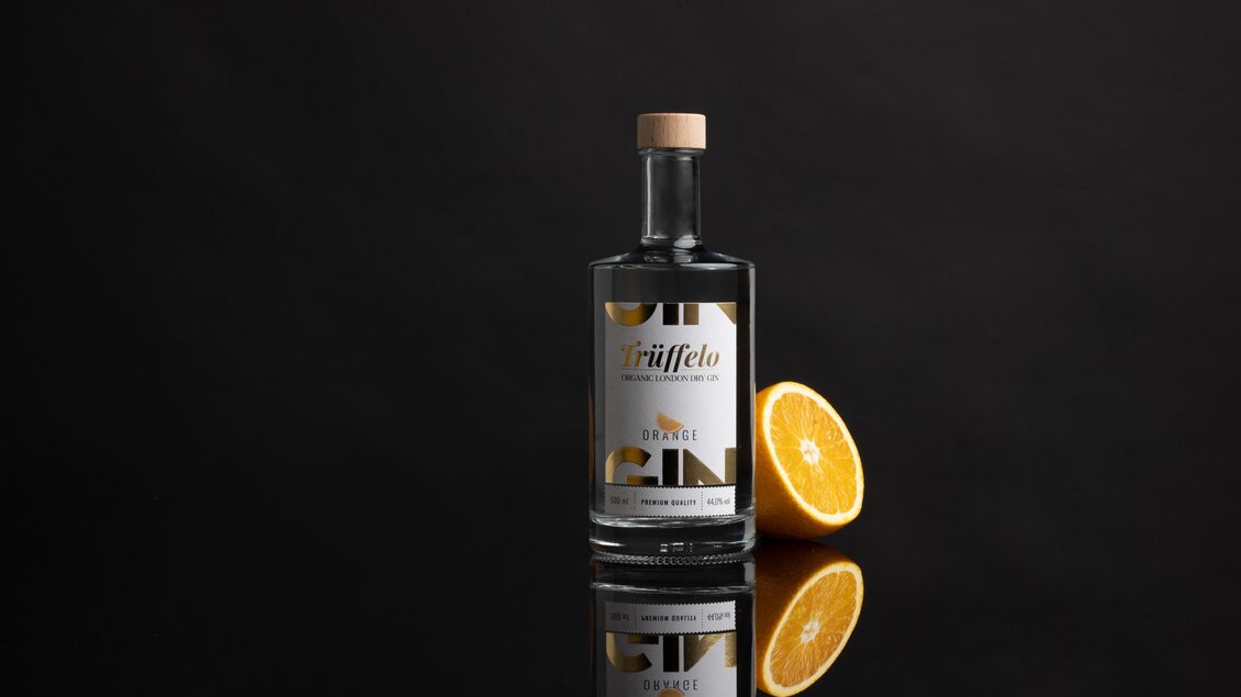 Eine stilvolle Flasche Gin steht auf einem schwarzen Hintergrund. Daneben liegt eine halbierte orange. | © True-fellow Handel GmbH