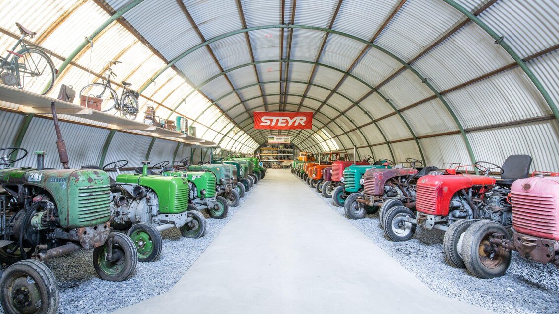 Eine Reihe von alten Traktoren in einer Lagerhalle. Die Traktoren sind in verschiedenen Farben angeordnet, und die Wände sind mit historischen Fahrrädern dekoriert. | © Steir. Rucksackdörfer-Schmid Philipp_Birgit Maier