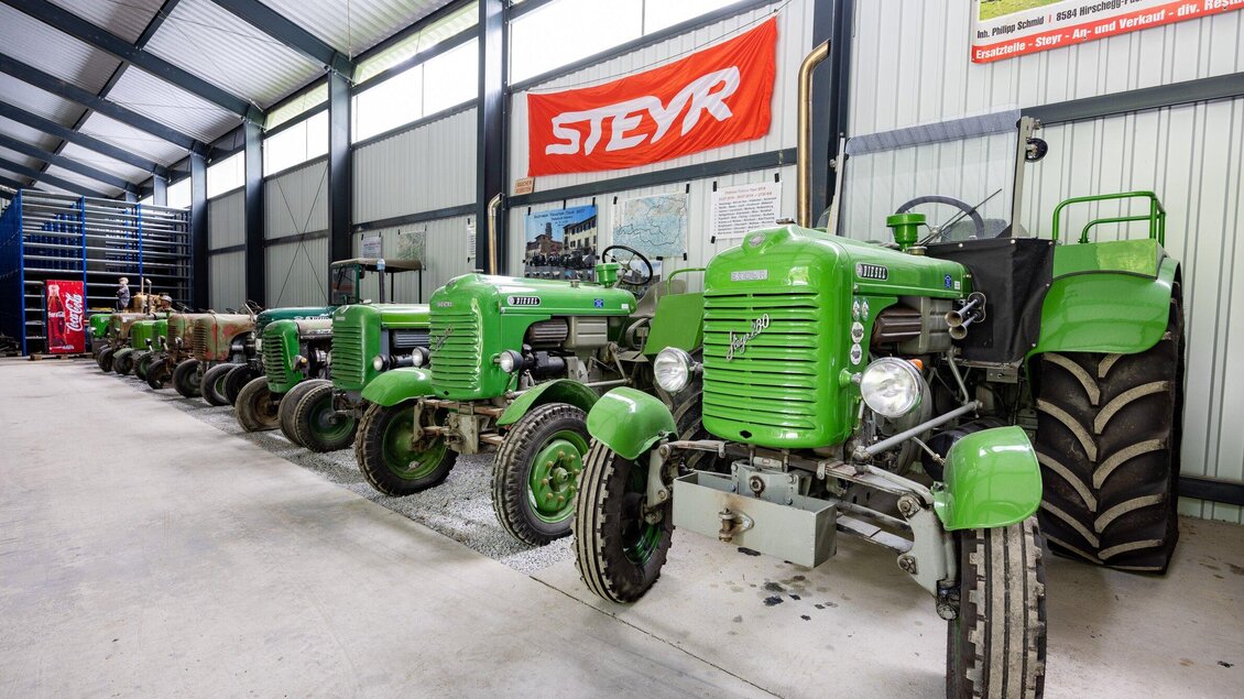 Eine Reihe von historischen Traktoren in einer Halle. Die Traktoren sind grün und stehen ordentlich aufgereiht, im Hintergrund ist ein Banner sichtbar. | © TV Region Graz-Harry Schiffer