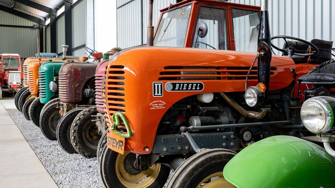 Eine Reihe von bunten Traktoren in einer Halle. Die Traktoren sind in verschiedenen Farben angeordnet, darunter orange und grün. | © TV Region Graz-Harry Schiffer