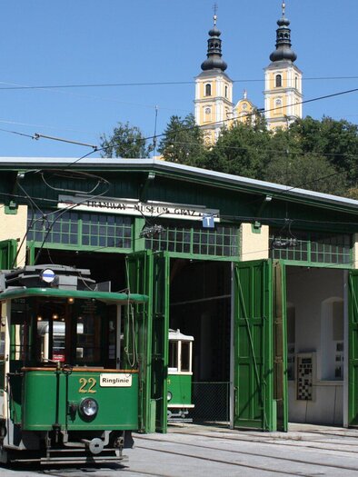 <p>Eine historische Straßenbahn vor einem Depot mit grünen Türen. Im Hintergrund sind Türme der Kirche Mariatrost sichtbar.</p> | © Tramway Museum Graz