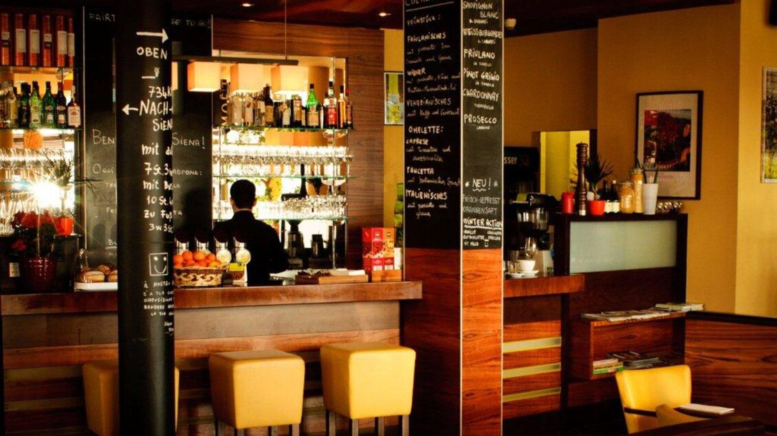 Eine stilvolle Bar mit einem Holztresen und Regalen voller Getränke. Im Hintergrund sind Tische und Stühle zu sehen, die eine gemütliche Atmosphäre schaffen. | © Trattoria Siena