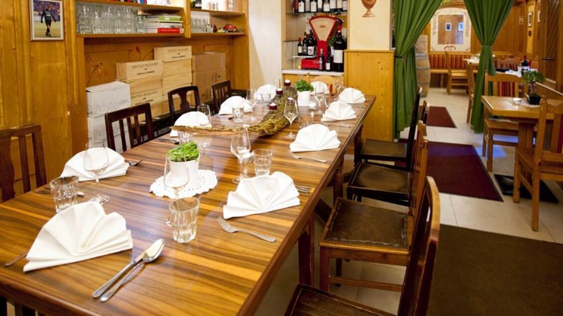 Ein einladendes Restaurant mit einem langen Holztisch, der für ein Essen gedeckt ist. Die Wände sind mit Weinflaschen und dekorativen Elementen geschmückt. | © Trattoria