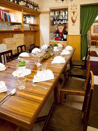 Ein einladendes Restaurant mit einem langen Holztisch, der für ein Essen gedeckt ist. Die Wände sind mit Weinflaschen und dekorativen Elementen geschmückt. | © Trattoria