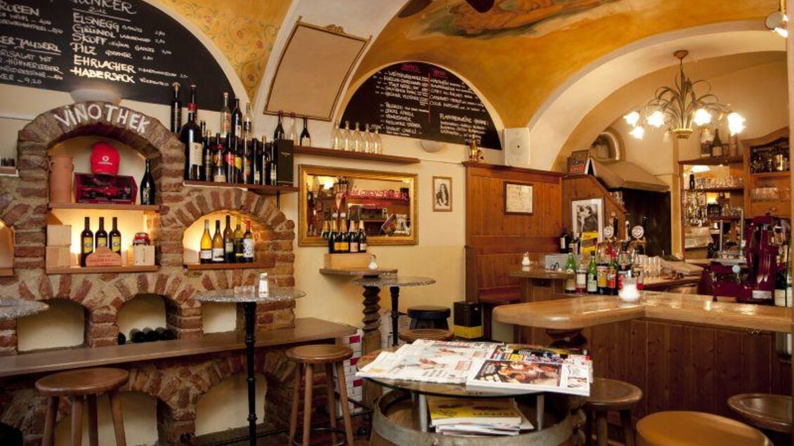 Eine gemütliche Weinbar mit Holzregalen und einer warmen Beleuchtung. Im Hintergrund sind eine Auswahl an Weinen und eine Tafel mit Speisen zu sehen. | © Trattoria