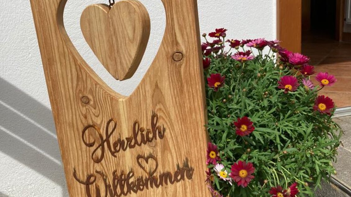 Ein rustikales Willkommensschild aus Holz mit einem herzförmigen Ausschnitt. Daneben blühen bunte Blumen in einem Topf.