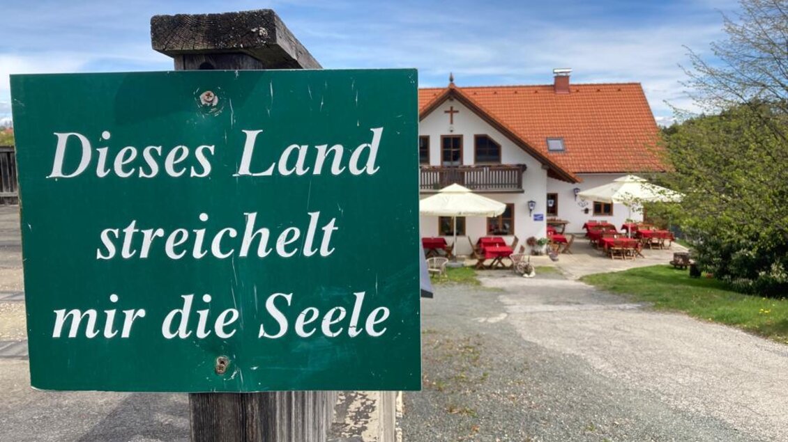 Ein Schild mit der Aufschrift „Dieses Land streichelt mir die Seele“ steht vor einem Gasthaus. Im Hintergrund sind Tische und Stühle im Freien zu sehen, umgeben von einer ruhigen Landschaft.