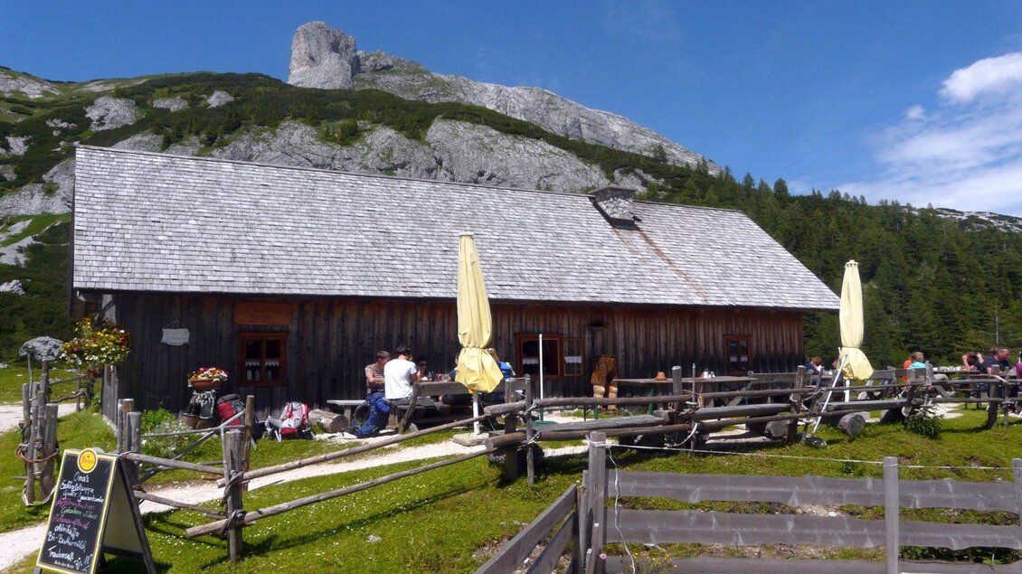 Eine gemütliche Berghütte mit einer Holzfassade, umgeben von Bergen und grünen Wiesen. Viele Menschen sitzen im Freien und genießen die Natur. | © TVB Ausseerland Salzkammergut/S. Zink