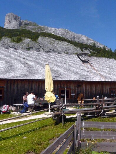 Eine gemütliche Berghütte mit einer Holzfassade, umgeben von Bergen und grünen Wiesen. Viele Menschen sitzen im Freien und genießen die Natur. | © TVB Ausseerland Salzkammergut/S. Zink