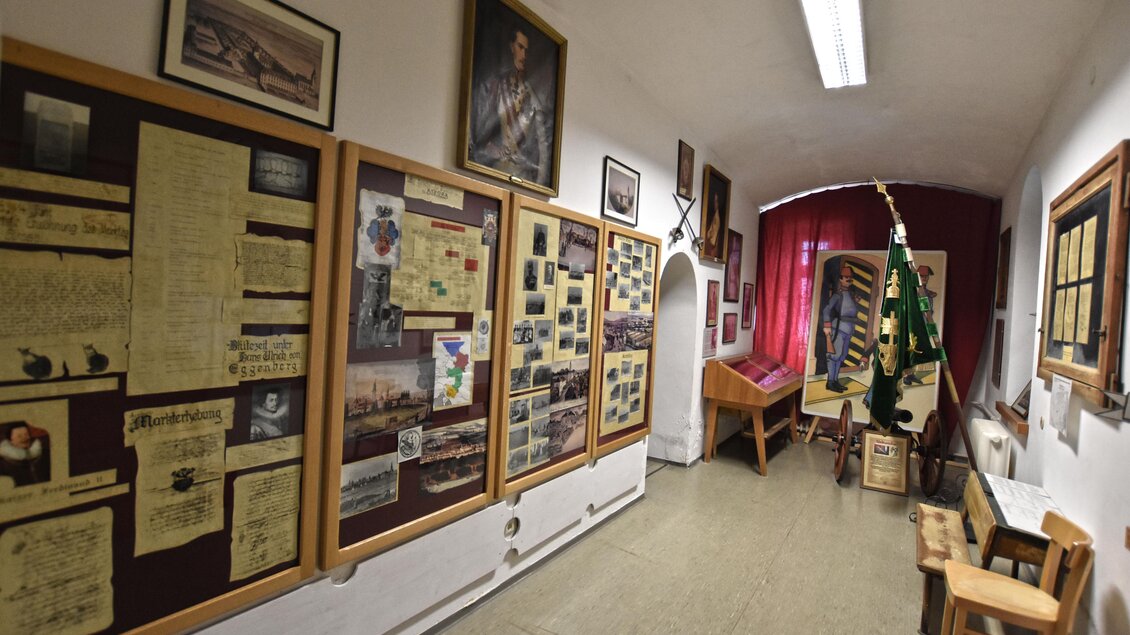 Ein langer Flur mit Wänden, die mit historischen Fotografien und Dokumenten dekoriert sind. Auf der rechten Seite stehen einige Möbelstücke und eine grüne Fahne. | © BMLV/Robert Gießauf