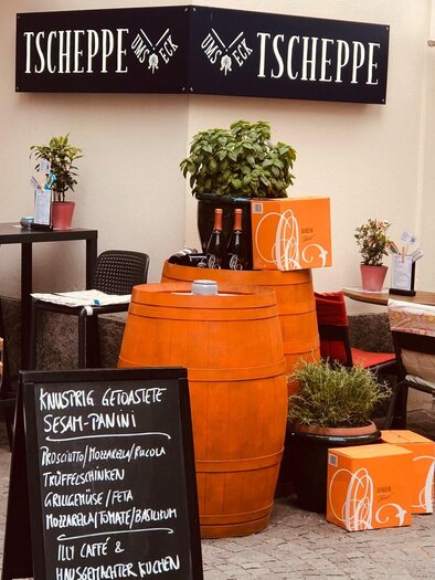 Ein einladendes Café mit Außenbereich, Tischen und Stühlen. Ein schwarzes Schild zeigt die Menüoptionen an, während Blumenkübel für eine gemütliche Atmosphäre sorgen. | © Tscheppe ums Eck