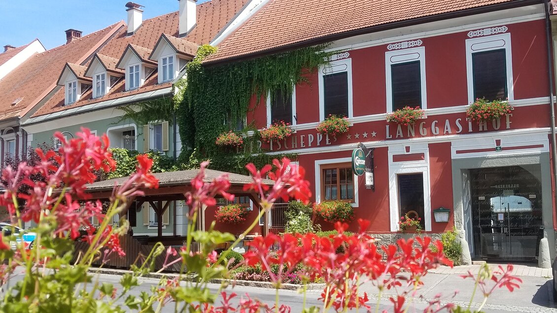 Ein charmantes Gasthaus mit roten Wänden und Blumenkästen. Im Vordergrund blühen bunte Blumen und im Hintergrund sind schöne, traditionelle Häuser zu sehen. | © Tscheppe