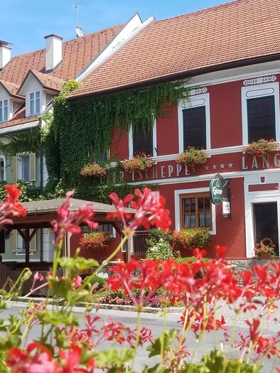 A charming inn with red walls and flower boxes. In the foreground, colorful flowers are blooming, and in the background, beautiful traditional houses can be seen. | © Tscheppe