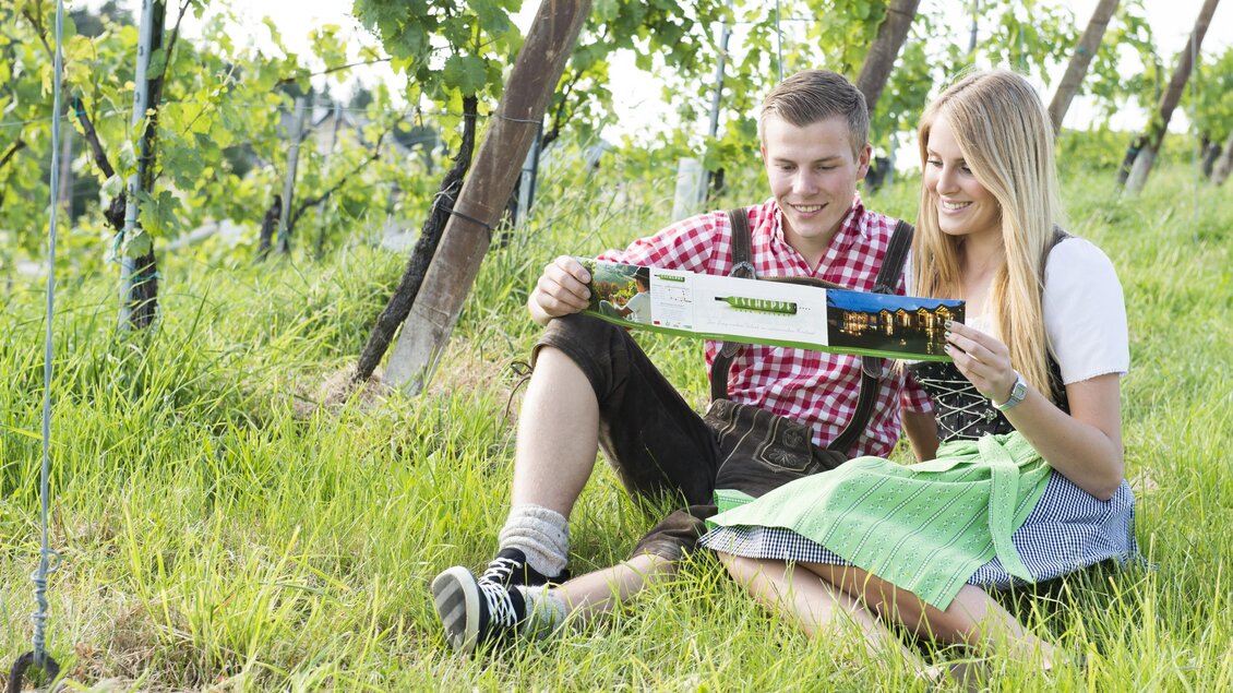 Ein Paar sitzt im Weinberg und schaut sich eine Broschüre an. Sie tragen traditionelle Trachten und die Umgebung ist grün und einladend. | © Tscheppe