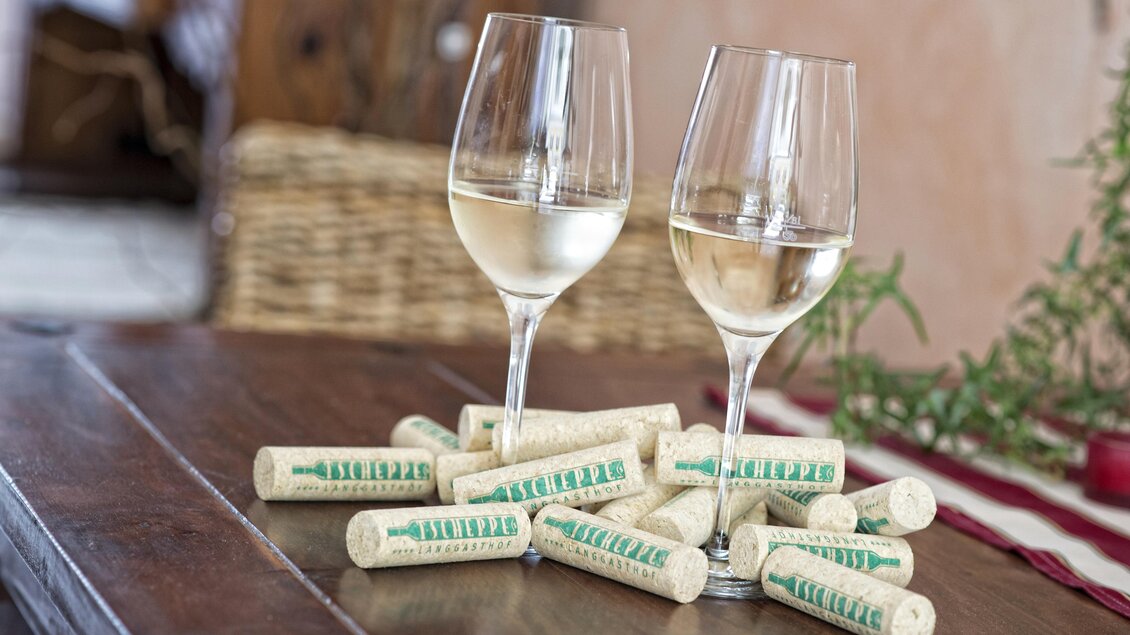 Zwei Gläser Weißwein stehen auf einem Holztisch, umgeben von Korken. Im Hintergrund sind dekorative Elemente zu sehen. | © Tscheppe