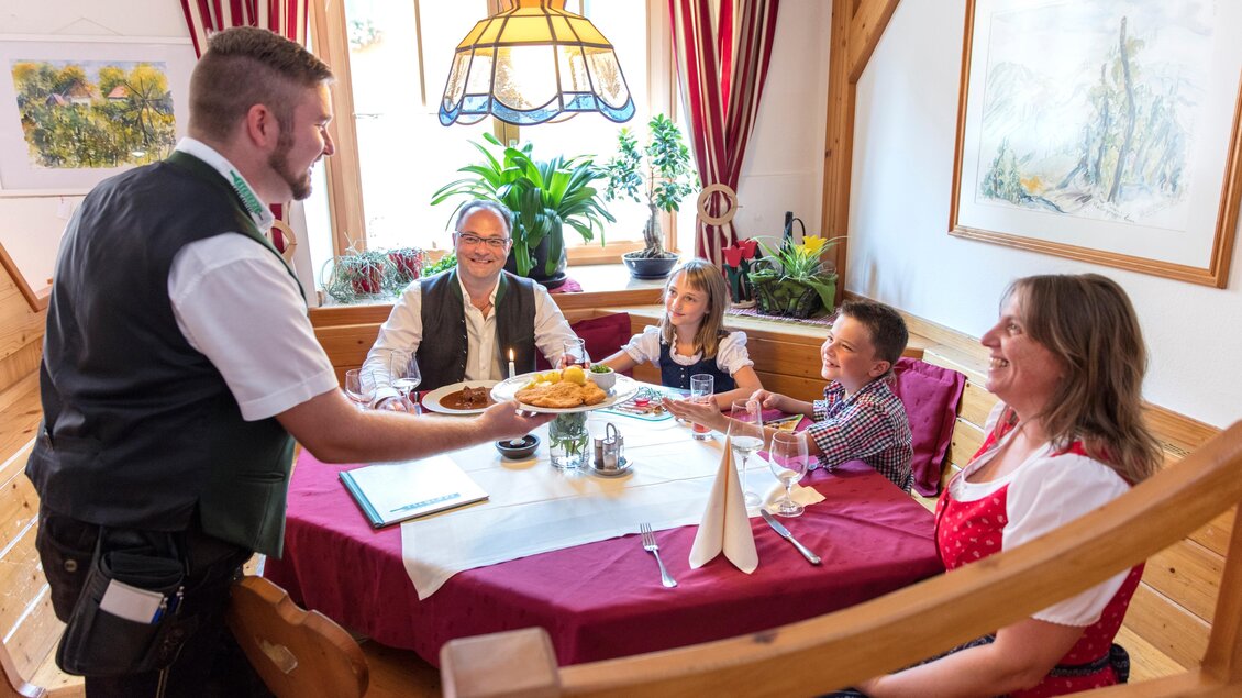 Eine Familie genießt ein Essen in einem gemütlichen Restaurant. Der Kellner serviert warme Speisen am Tisch. | © Tscheppe
