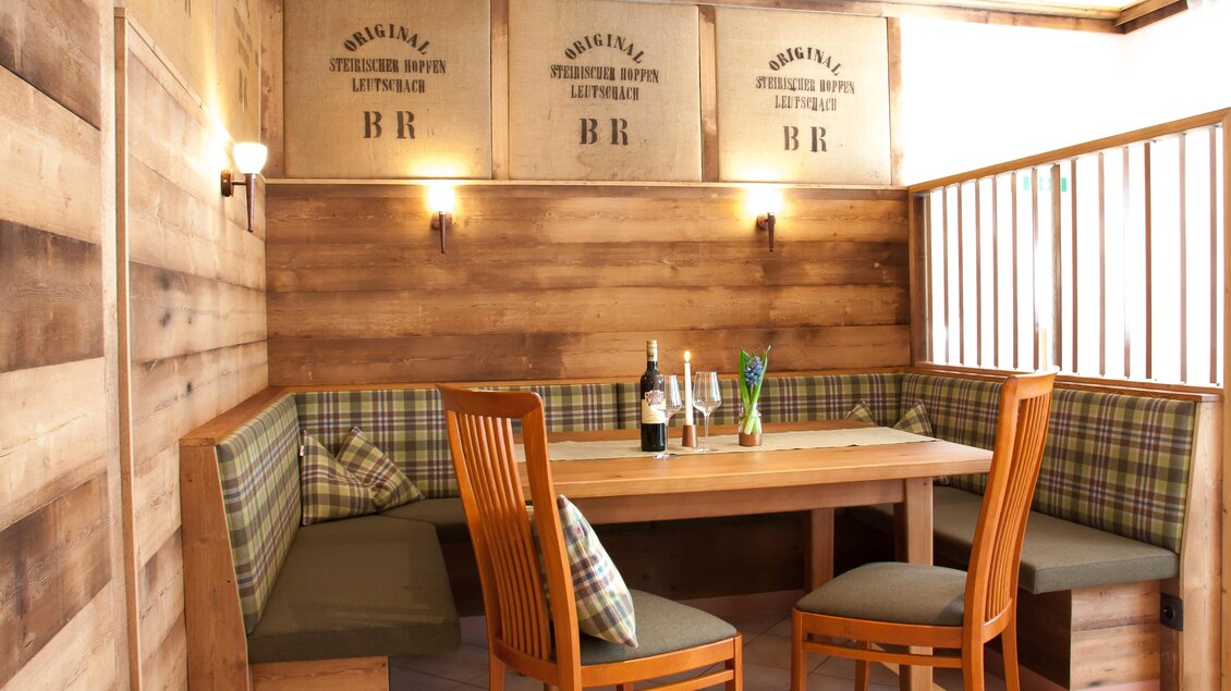 Ein gemütlicher Essbereich mit Holzvertäfelung und einem großen Tisch. Die Sitzecke ist mit Kissen ausgestattet und es stehen Weinflaschen auf dem Tisch. | © Tscheppe