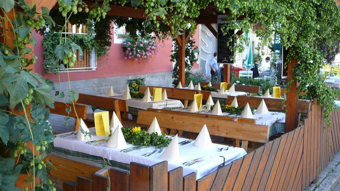 Ein einladendes Restaurant mit Tischen im Freien, geschmückt mit grünen Pflanzen. Die Tische sind elegant gedeckt und bereit für Gäste. | © Tscheppe