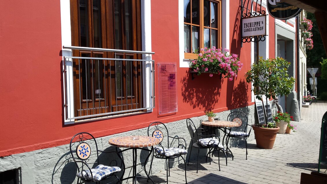 Ein eleganter Außenbereich mit kleinen Tischen und Stühlen vor einem roten Gebäude. Bunte Blumen und ein schattenspendender Baum schaffen eine einladende Atmosphäre. | © Tscheppe