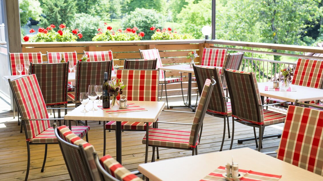 Eine einladende Terrasse mit Tischen und Stühlen in einem Restaurant. Die Möbel sind mit rot-karierten Kissen dekoriert und die Umgebung ist grün und blühend. | © Tscheppe